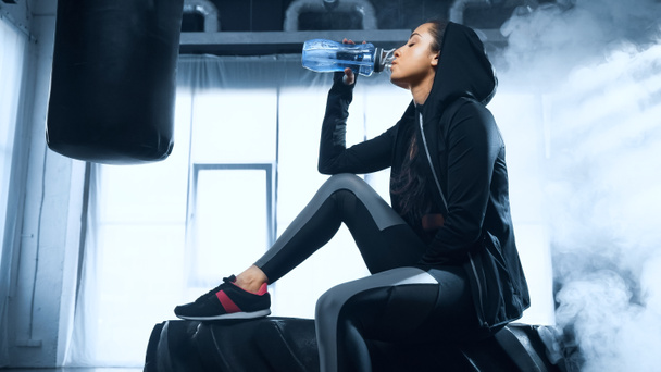 sportvrouw in hoodie zittend op band en drinkwater uit sportfles - Foto, afbeelding
