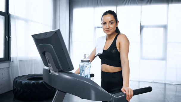 Lächelnde Sportlerin mit Handtuch auf Laufband neben Sportflasche mit Wasser im Fitnessstudio - Foto, Bild