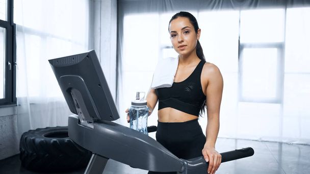sportswoman walking on treadmill and reaching sports bottle with water in gym - Фото, зображення