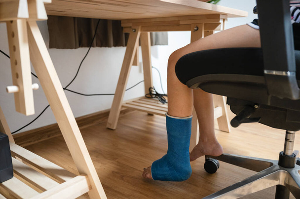 Травмированная женщина со сломанной ногой в гипсе, работающая за деревянным столом - Фото, изображение