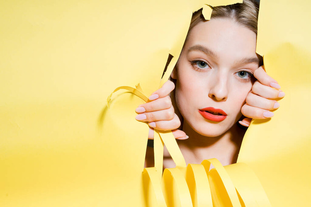 The face of a girl with bright makeup looking through a hole in a yellow background, pushing it apart with her hands - 写真・画像