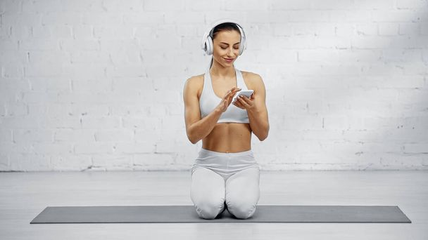Femme sportive souriante écoutant de la musique dans un casque et utilisant un téléphone portable sur un tapis de fitness  - Photo, image