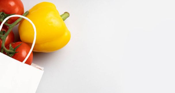 Bolsa de papel blanco con tomates y pimienta amarilla sobre fondo gris, lugar para texto, vista superior. - Foto, Imagen