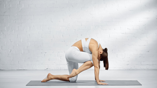 Seitenansicht einer Frau in weißer Sportbekleidung, die sich auf einer Yogamatte beugt  - Foto, Bild