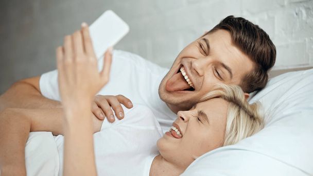 幸せな若いカップルが舌を出しながらスマホで自撮り  - 写真・画像
