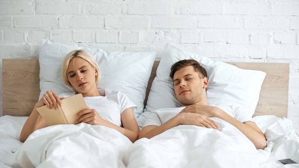 young blonde woman reading book near sleeping boyfriend in bedroom  - 写真・画像