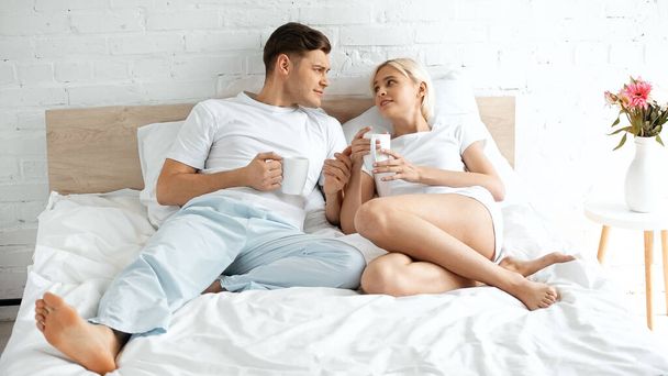 young couple looking at each other while holding cups of coffee in bed - Foto, Imagem