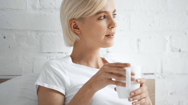 ブロンド女性で白いtシャツを保持カップのコーヒーでベッドルーム - 写真・画像