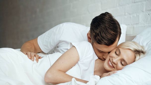 happy man kissing smiling woman lying with closed eyes in bed - Фото, изображение