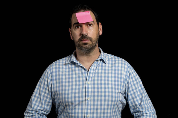 Ein bärtiger Mann in einem weiß-blau karierten Hemd mit einem Papier auf der Stirn, das isoliert auf schwarzem Studiohintergrund klebt. Spaßkonzept - Foto, Bild