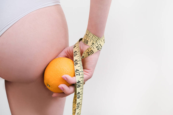 young woman is holding an orange and a measuring tape on a light background. Cellulite problem concept - Zdjęcie, obraz