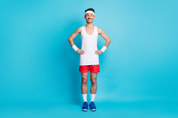 Full length photo of sporty man hold hand waist wear white singlet gumshoes isolated over blue color background - Foto, Imagen