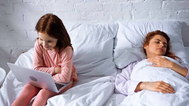 Kid using laptop while mother sleeping on bed  - Photo, Image