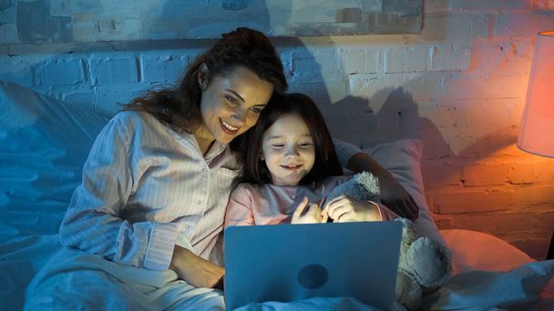Χαμογελαστή γυναίκα και κόρη που χρησιμοποιούν φορητό υπολογιστή στο κρεβάτι κατά τη διάρκεια της νύχτας  - Φωτογραφία, εικόνα