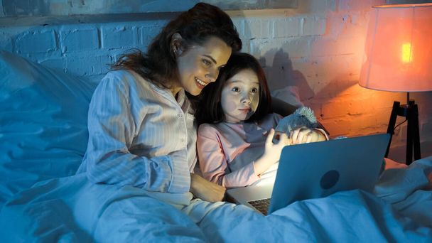 Mère souriante regardant ordinateur portable près de l'enfant avec ours en peluche sur le lit  - Photo, image