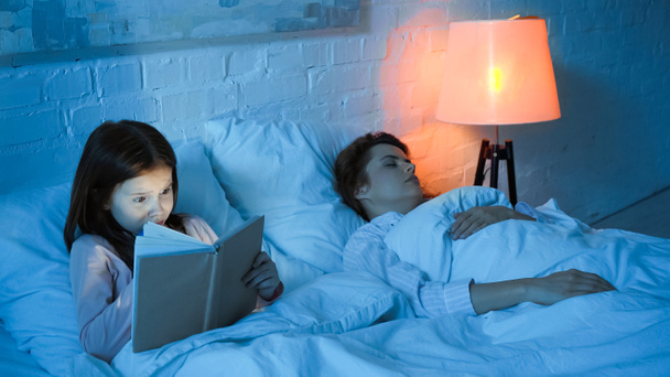Kid leitura livro perto de mãe dormindo na cama durante a noite  - Foto, Imagem