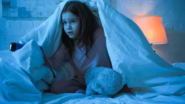 Испуганный ребенок держит фонарик рядом с плюшевым мишкой под одеялом на кровати  - Фото, изображение