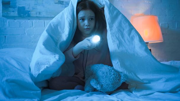 Chica sosteniendo linterna cerca de juguete suave en la cama en la noche  - Foto, Imagen
