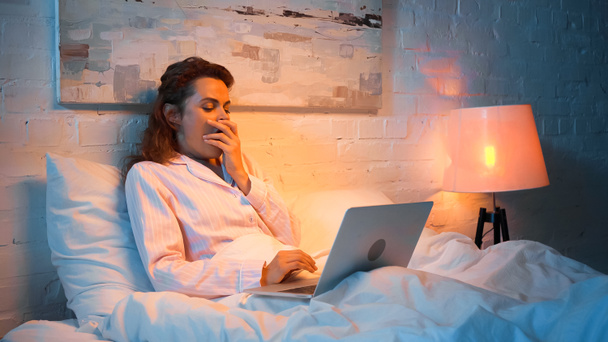 Kobieta w piżamie ziewanie podczas korzystania z laptopa na łóżku wieczorem  - Zdjęcie, obraz
