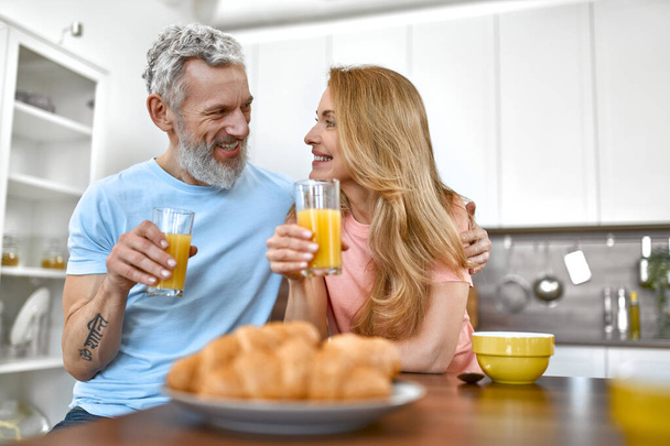 Ältere Paare frühstücken frühmorgens in der Küche und amüsieren sich. Ein Ehepaar frühstückt mit Saft und Croissants. - Foto, Bild