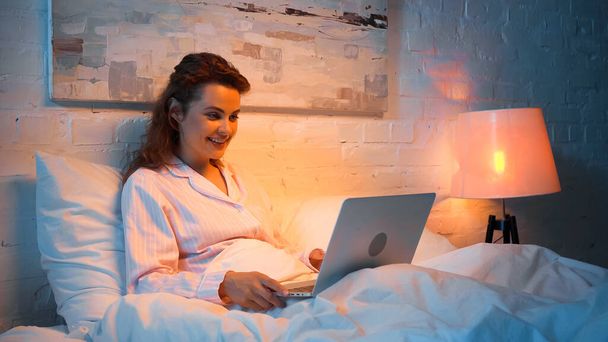 Yatakta dizüstü bilgisayar kullanan pijamalı neşeli bir kadın.  - Fotoğraf, Görsel