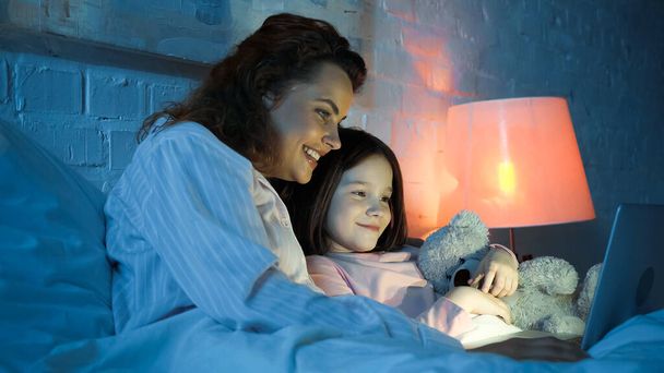 Iloinen nainen käyttää kannettavaa tietokonetta lähellä hymyilevä tytär pehmeä lelu sängyssä yöllä  - Valokuva, kuva