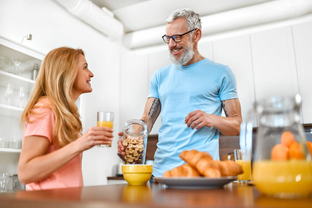 La pareja mayor está desayunando. Un hombre maduro vierte cereal en su plato y tiene un tiempo agradable con su esposa en la cocina por la mañana. - Foto, Imagen