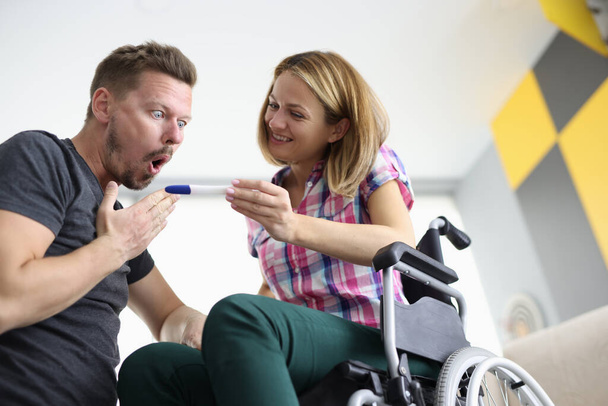 Радостная женщина в инвалидной коляске показывает удивленный тест на беременность мужчины - Фото, изображение