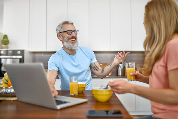 Ευτυχισμένο παντρεμένο ζευγάρι τρώνε πρωινό και διασκεδάζουν χρησιμοποιώντας το laptop στην κουζίνα. Υγιεινός τρόπος ζωής. - Φωτογραφία, εικόνα