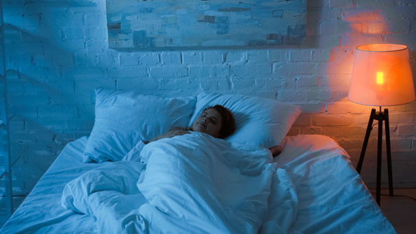 Kobieta śpi na łóżku w pobliżu lampy podłogowej w sypialni  - Zdjęcie, obraz