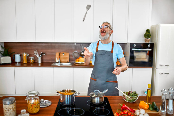 A mature elderly man in an apron prepares pasta in the kitchen. Vegan, vegetarian, healthy lifestyle. - Fotó, kép
