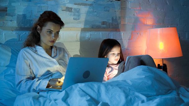Freiberuflerin nutzt Laptop neben Tochter mit digitalem Tablet auf Bett  - Foto, Bild