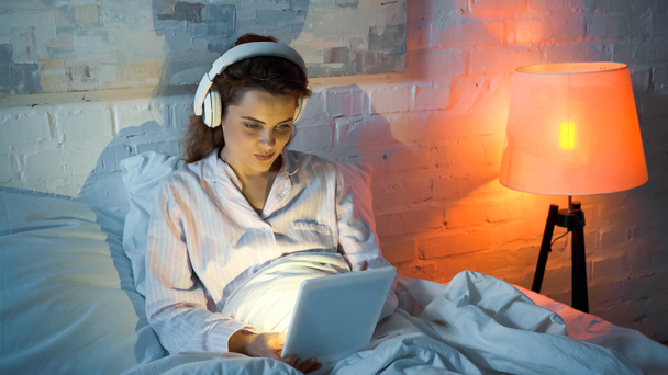 Γυναίκα σε ακουστικά χρησιμοποιώντας ψηφιακό δισκίο στο κρεβάτι κατά τη διάρκεια του βραδινού  - Φωτογραφία, εικόνα
