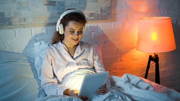 Uśmiechnięta kobieta korzystająca z tabletu cyfrowego i słuchawek w sypialni  - Zdjęcie, obraz