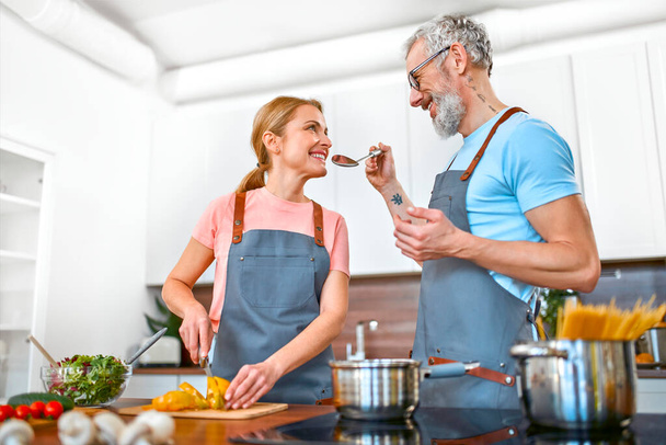 エプロンで幸せな高齢者のカップルは、キッチンでパスタや新鮮なサラダを準備しており、楽しい時間を過ごしています。ビーガン,ベジタリアン,健康的なライフスタイルのコンセプト. - 写真・画像