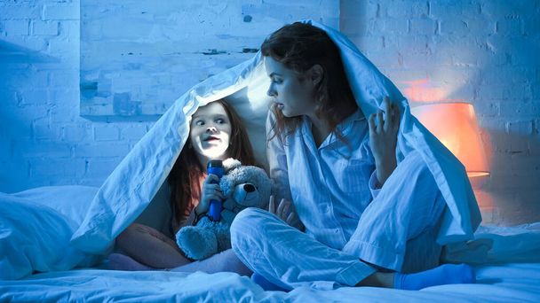 Kid with flashlight grimacing near mother and soft toy in bedroom  - Photo, Image