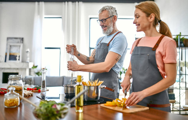 A boldog idős pár kötényben tésztát és friss salátát készít a konyhában, és jól érzik magukat. Vegán, vegetáriánus, egészséges életmód koncepció. - Fotó, kép