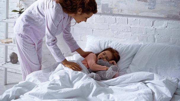 ベッドの上で寝ている子供の近くに立つパジャマの女性  - 写真・画像