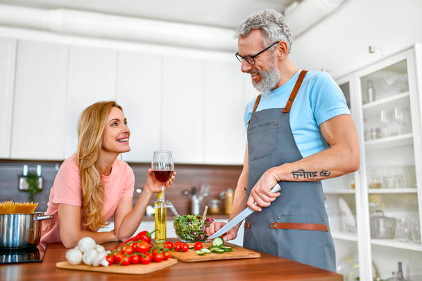 Glückliches Seniorenpaar in der Küche. Reifer Mann in Schürze bereitet romantisches Abendessen für seine Frau vor. Veganes, vegetarisches, gesundes Lebensstilkonzept. - Foto, Bild