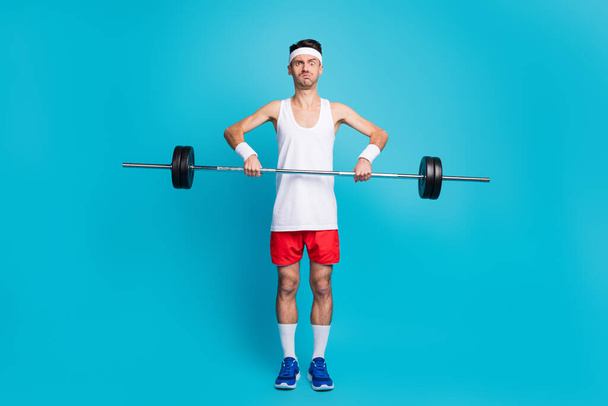 Full size photo of young man pouted cheekbones lift barbells heavy exercise bodybuilding isolated over blue color background - Фото, изображение