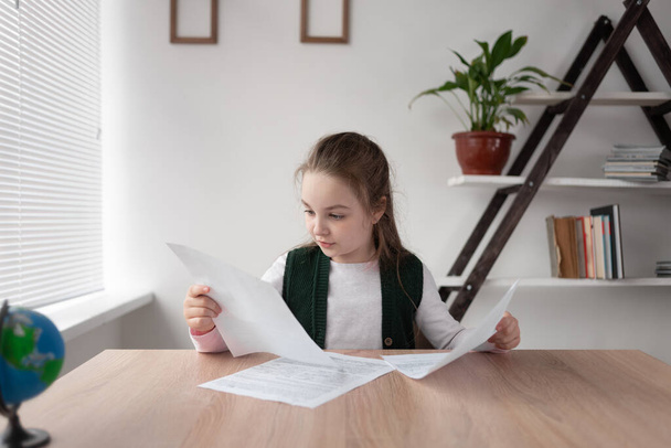 Schoolgirl student girl sits at home and learns online distance. Pupil is holding sheets of paper with homework in his hands and thinking intently over the answers to the teacher via the video call - Foto, immagini