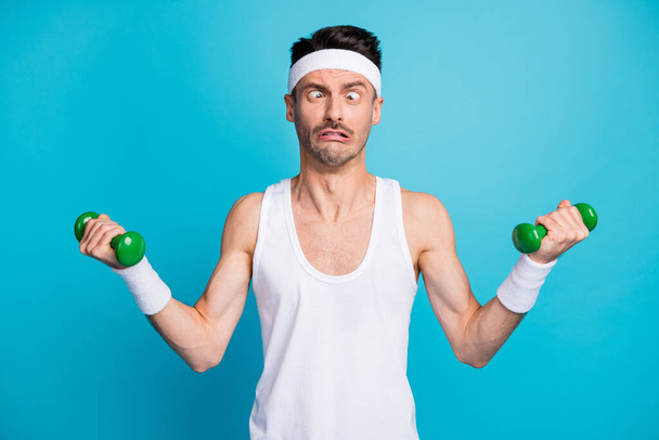 Photo of young sportsman train exercise with heavy dumbells grimacing eyes look different way isolated over blue color background - Fotografie, Obrázek