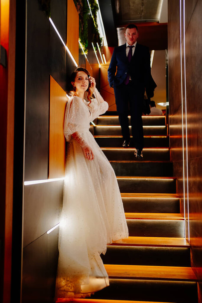 The groom in a suit and the bride in a white dress stand on poorly lit staircase - Foto, imagen