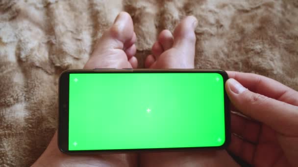 Egy férfi ül egy bolyhos takarón, és tart egy okostelefont zöld képernyővel a lábán. Bőrkeményedések a lábujjakon.A telefon nézete felülről. - Felvétel, videó