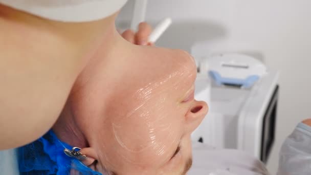 Косметолог наносит прозрачную гелевую маску на лицо женщины старшего возраста кистью. Пожилая клиентка получает антивозрастную процедуру. Вертикальное видео. уход за кожей лица и шеи в спа. 4 k видео - Кадры, видео