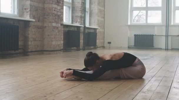 Pełen strzał atrakcyjnej młodej baleriny przygotowującej się do tańca baletowego wykonującej ćwiczenia rozciągające siedzącej na drewnianej podłodze w przestronnym studio tanecznym - Materiał filmowy, wideo