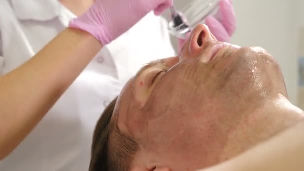 Kosmetologia, zabiegi kosmetyczne dla mężczyzn. Kosmetyk używa ciemnej maski do twarzy ze szczotką. Mężczyzna leżący na kanapie i mający zabieg pielęgnacji twarzy, w klinice kosmetycznej, dermatologii. 4 tys. wideo - Materiał filmowy, wideo