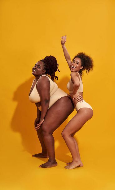 Full length shot of two funny slim and plus size african american women in beige underwear looking joyful while posing isolated over yellow background - Foto, Imagem