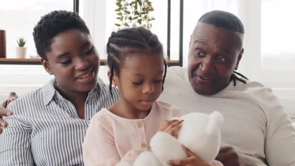Happy African family sitting on sofa, mom daughter and grandfather hugging chatting at home, mature man father shows pigtails braids to little child black kid girl, talks about similarity hairstyles - Footage, Video