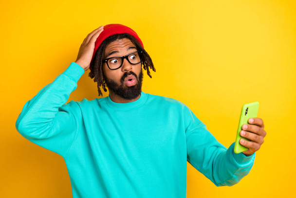 Portrait of brunette impressed guy hold telephone wear cap spectacles blue pullover isolated on yellow vivid color background - Fotoğraf, Görsel
